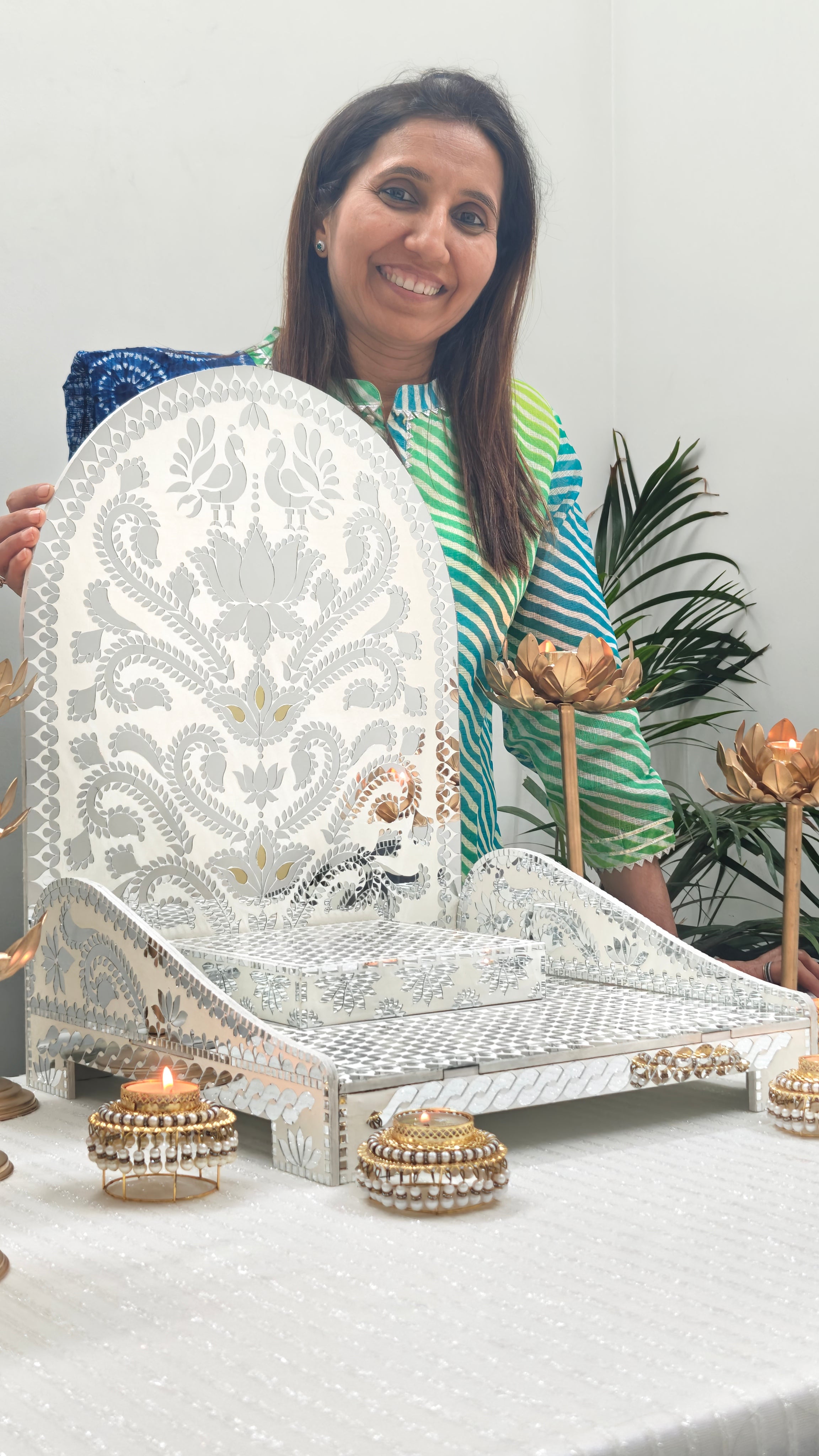 Woman showcasing decorative white mirror mosaic chowki with floral bird design, elegant pooja setup with lotus candle holders for festive mandir and home décor.