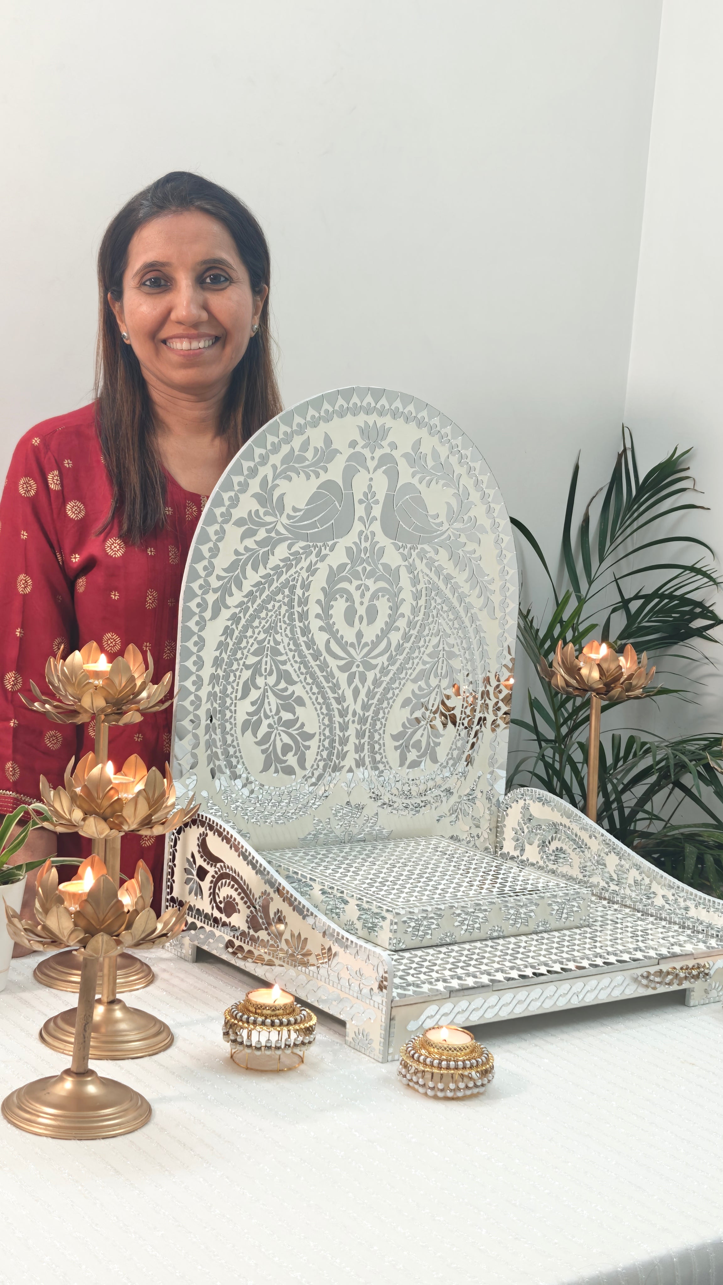 Woman showcasing decorative white mirror mosaic chowki with peacock floral design, elegant pooja setup with lotus candle holders for festive home temple décor.