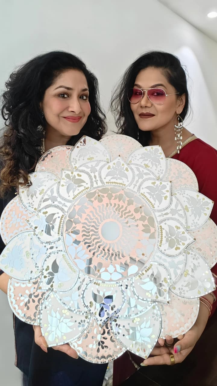 Two women holding a large decorative MDF rangoli base with intricate floral and mirror-cut patterns, showcasing a finished Lippan art design.